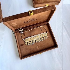 May include: A gold-toned necklace with intricate detailing, displayed inside an open brown velvet jewelry box. The necklace features a central design with dangling elements and is presented against a brown interior. The box has a gold clasp and the text "JEWELS by Layyan".