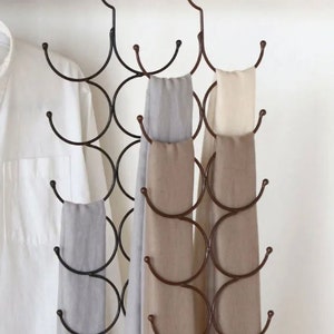 May include: A bronze metal scarf organizer with multiple circular hooks, holding several scarves in shades of gray and beige. A white shirt hangs on a wooden rod in the background. The organizer is designed to hang in a closet.