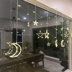 May include: A window with a string of lights featuring stars and crescent moons. The lights are hung on a white wall and reflect off a wooden table and chairs in the background.
