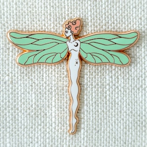 Puede incluir: Un pin de esmalte dorado con un diseño de libélula estilizada con un cuerpo humano y alas verdes. La libélula tiene una luna creciente en el pecho y una pequeña cruz en el abdomen.