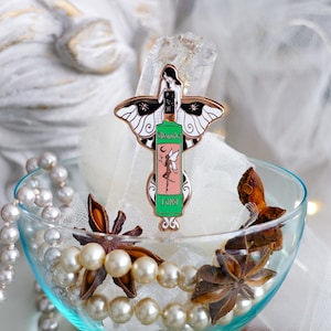 Pode incluir: Um broche de esmalte verde e rosa com uma fada com asas e uma garrafa de absinto. A garrafa está etiquetada como "Absinthe Luna". O broche está em uma tigela de vidro com anis estrelado e pérolas.