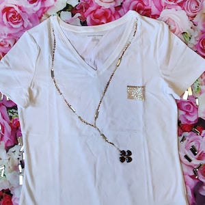 Könnte beinhalten: Weißes T-Shirt mit V-Ausschnitt, einer glitzernden Tasche und einer braunen und silbernen Perlenkette mit einem Blumenanhänger.