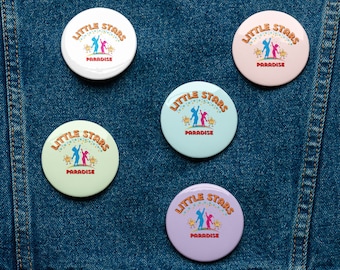 Può includere: Cinque bottoni rotondi su uno sfondo di denim blu chiaro. Ogni bottone ha uno sfondo bianco con una grafica colorata di due bambini che ballano con il testo "Little Stars Paradise" in arancione.