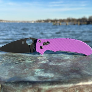 May include: A purple and black Spyderco knife with a carbon fiber handle and a CPM REX 45 blade.