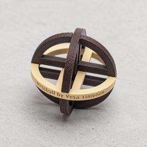 May include: A spherical puzzle composed of interlocking dark brown wooden rings and a light beige central band. The beige band features the engraved text Metaball by Vesa Timonen. The object consists of multiple intersecting circular components.