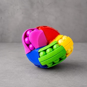 May include: A colorful puzzle ball with sections in red, yellow, green, blue, and pink. Each section holds smaller, brightly colored spheres. The toy is designed to be manipulated and solved, offering a tactile and visual challenge.