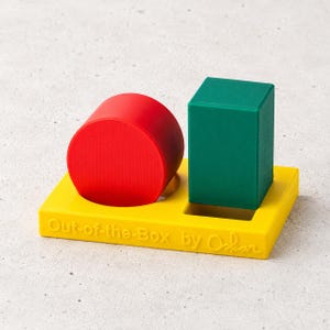 Puede incluir: Una base rectangular amarilla con el texto Out-of-the-Box by Oskar grabado en el frente. La base presenta dos recortes que sostienen un cilindro rojo y un prisma rectangular verde. Los objetos están dispuestos uno al lado del otro.