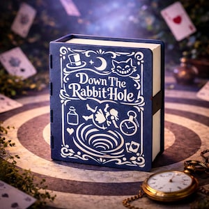 Puede incluir: Una caja azul con forma de libro con las palabras "Down the Rabbit Hole" en escritura blanca. La portada presenta ilustraciones de un sombrero, un gato y otras imágenes de Alicia en el país de las maravillas. Un reloj de bolsillo dorado con una cadena descansa cerca.