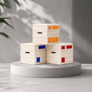 Op de afbeelding: Drie beige, stapelbare opbergdozen met gekleurde accentpanelen in oranje, blauw en rood. De dozen zijn gerangschikt op een grijze, cirkelvormige sokkel. Elke doos heeft een kleine handgreep.