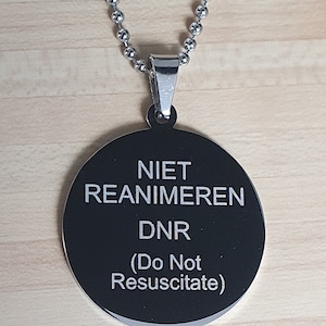Könnte beinhalten: Ein schwarzer, kreisförmiger Anhänger mit dem Text "NIET REANIMEREN DNR (Do Not Resuscitate)" in weißer Schrift. Der Anhänger hängt an einer silbernen Kugelkette.