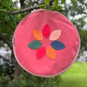 May include: A round pink bag with a zip fastening and a flower design made of colourful leather patches.