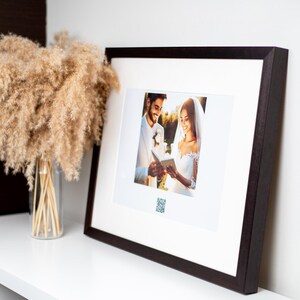 Könnte beinhalten: Ein schwarzer gerahmtes Foto von einem Paar in Hochzeitskleidung. Die Braut und der Bräutigam lächeln und schauen sich an. Das Foto steht auf einem weißen Regal mit einer Vase mit getrocknetem Pampasgras.