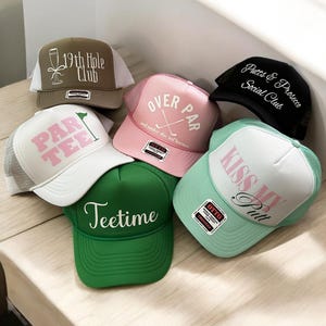 May include: Six different trucker hats with various golf-themed designs. The hats are in different colors, including green, pink, white, black, and brown. The designs include "19th Hole Club", "Par Tee", "Over Par", "Putts & Prosecco Social Club", "Kiss My Putt", and "Teetime".