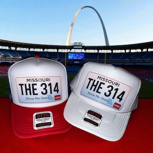 May include: Two trucker hats with a white front panel and a red or white brim. Each hat features a license plate design with "MISSOURI THE 314 Show Me State" printed on it. The background shows a stadium and the Gateway Arch.