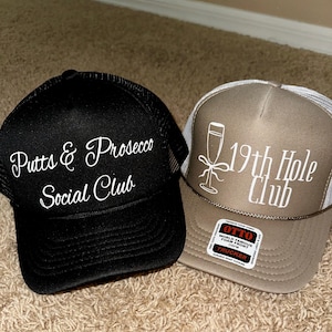 May include: Two black and brown trucker hats with white embroidered text. The black hat says "Putts & Prosecco Social Club". The brown hat says "19th Hole Club" and has a white glass of champagne graphic.