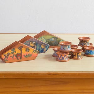 May include: A collection of terracotta napkin holders and napkin rings. The napkin holders feature hand-painted animal and floral designs in orange, green, and blue. The napkin rings have similar patterns.