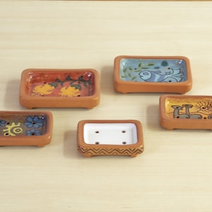 May include: A collection of five small, rectangular ceramic soap dishes. Each dish has a terracotta-colored exterior and a painted interior with various designs, including flowers, fish, and abstract patterns. The dishes are displayed on a light-colored wooden surface.