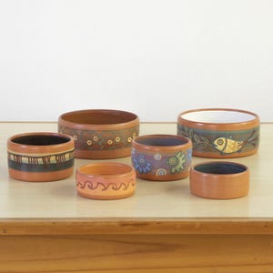 May include: A collection of six handcrafted ceramic bowls in various sizes. The bowls feature earthy tones of brown and orange, with decorative patterns including geometric shapes, swirls, and a fish design. The bowls are displayed on a light-colored wooden surface.