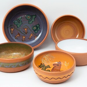 May include: A collection of handmade ceramic bowls in various sizes and designs. The bowls feature earthy tones with painted interior designs, including floral and abstract patterns. The rims are a natural terracotta color.