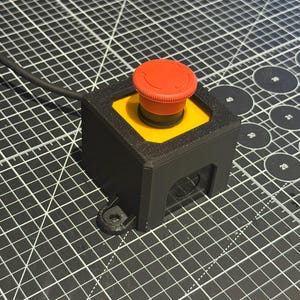 May include: A black and yellow emergency stop button with a red button top. The button is mounted on a black square base with a black cable attached. A small black piece is next to the base.