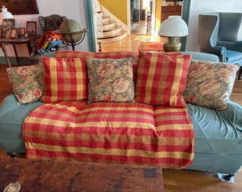 May include: A light blue sofa covered with a red and yellow plaid throw blanket and five decorative cushions. Three cushions feature a red and yellow plaid pattern, while two cushions display a floral print with pink and green tones.