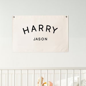 May include: A cream-colored banner with the names "HARRY" and "JASON" in black lettering hangs above a white crib. The crib contains a mattress, pillows, and a stuffed animal. A white bunny-shaped lamp sits on the floor.