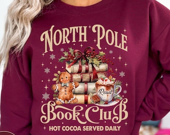 May include: Maroon sweatshirt with a festive design. The graphic reads "North Pole Book Club" with a gingerbread man, books, and a mug of hot cocoa. The text "Hot Cocoa Served Daily" is also included.