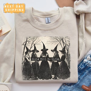 May include: A beige sweatshirt with a vintage illustration of four witches in black dresses and pointed hats. The witches are standing in a circle in a forest setting with trees and a full moon. The text "Girls Will Be Girls." is printed below the illustration.