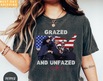 May include: A dark grey t-shirt with a graphic of a group of people in front of a US flag. The text "Grazed and Unfazed" is printed below the graphic.