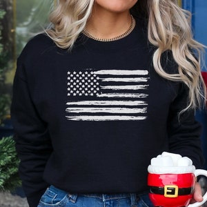 May include: A navy blue sweatshirt featuring a distressed white American flag design. The sweatshirt has a crew neck and long sleeves. The image also shows a Santa-themed mug filled with a white substance.