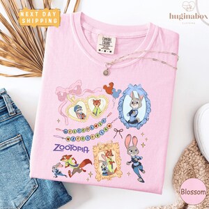 a pink t-shirt with various cartoon animal characters and a heart-shaped design, along with a charm and a tag. The t-shirt is placed on a white surface, and there are some jeans and a bracelet visible in the background.