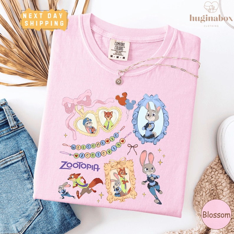 a pink t-shirt with various cartoon animal characters and a heart-shaped design, along with a charm and a tag. The t-shirt is placed on a white surface, and there are some jeans and a bracelet visible in the background.