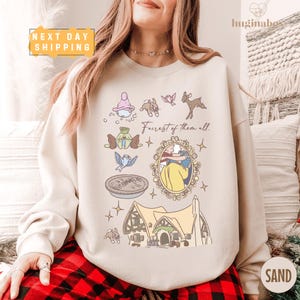 May include: Sand-colored sweatshirt featuring a whimsical illustration with the text "Fairest of them all." The design includes cartoon images of woodland creatures, a cottage, and a framed image of a princess. The sweatshirt has a crew neck.