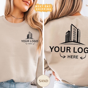 May include: A sand-colored crewneck sweatshirt featuring a black graphic of a city skyline on the front left chest and a larger version of the same skyline on the back. Both graphics include the text YOUR LOGO HERE with arrows pointing inward.