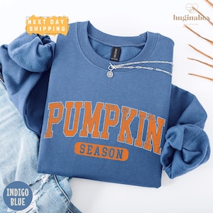 A blue sweatshirt with the word "PUMPKIN" printed in orange letters, along with the word "SEASON" below it, is displayed on a white background.