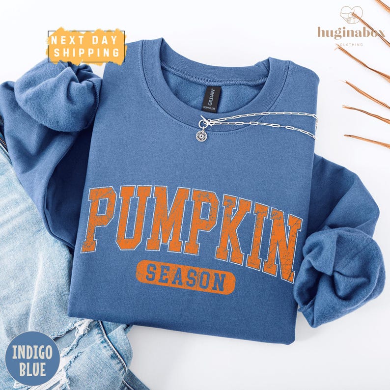A blue sweatshirt with the word "PUMPKIN" printed in orange letters, along with the word "SEASON" below it, is displayed on a white background.