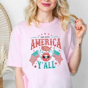 May include: Pale pink t-shirt with a patriotic design. The graphic includes the word "AMERICA" in red, the text "LAND OF THE FREE", and two American flags. The design also features the text "Y'ALL" in teal and the year "EST. 1776".