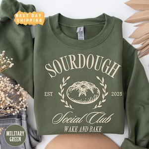 May include: A green sweatshirt with a white graphic design that says "Sourdough Social Club Est 2025 Wake and Bake" with a drawing of a loaf of bread.
