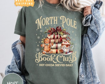 May include: Green t-shirt with a Christmas-themed design featuring a gingerbread man, a stack of books, a mug of hot cocoa, and the text "North Pole Book Club + Hot Cocoa Served Daily".