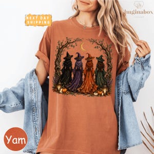 A woman wearing a t-shirt with a graphic design of four women dressed in witch costumes, surrounded by autumn elements such as pumpkins and a crescent moon.