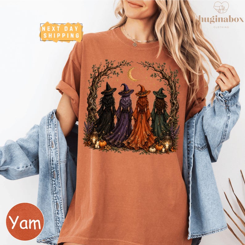 A woman wearing a t-shirt with a graphic design of four women dressed in witch costumes, surrounded by autumn elements such as pumpkins and a crescent moon.