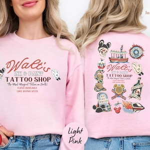 May include: Light pink sweatshirt featuring the text "Walt's Ink & Paint Tattoo Shop" with various tattoo-style illustrations. The design includes a skull with Mickey Mouse ears, a boat, a snake, and other themed graphics. The back of the sweatshirt also has a similar design.