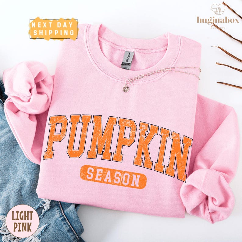 A pink sweatshirt with the word "PUMPKIN" printed on it, along with the word "SEASON" below it. The sweatshirt is folded and placed on a white background, with a pair of jeans visible in the background.