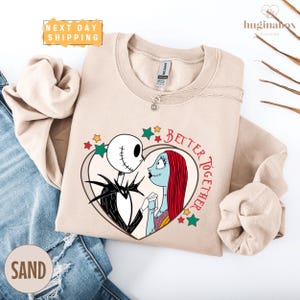 A beige sweatshirt with a graphic design featuring the characters Jack Skellington and Sally from the movie "The Nightmare Before Christmas". The design includes a heart shape with the text "Better together" and stars surrounding the characters.