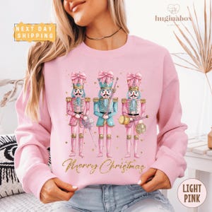 Puede incluir: Sudadera rosa claro con un diseño de acuarela de tres cascanueces con uniformes rosas y turquesas, sosteniendo varios instrumentos. Las palabras "Merry Christmas" están escritas en letras doradas debajo de los cascanueces. La sudadera es de color rosa claro.
