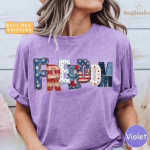 May include: A violet t-shirt with the word "FREEDOM" appliquéd across the chest. The letters are in a patchwork style, incorporating red, white, and blue fabrics with star and stripe patterns. The shirt has rolled-up sleeves.