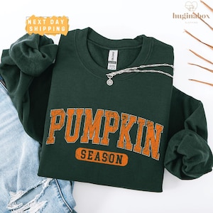 A green sweatshirt with the word "PUMPKIN" printed in orange letters, along with the word "SEASON" below it, is displayed on a white background.