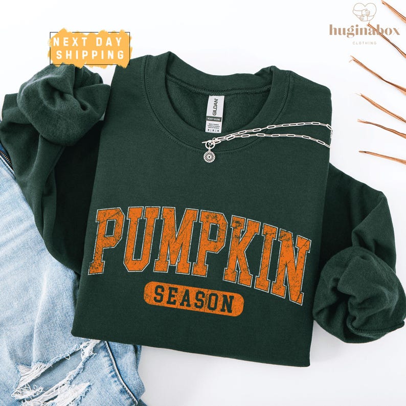 A green sweatshirt with the word "PUMPKIN" printed in orange letters, along with the word "SEASON" below it, is displayed on a white background.