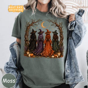 A person wearing a green t-shirt with a Halloween-themed graphic design featuring four witch figures in a forest setting.