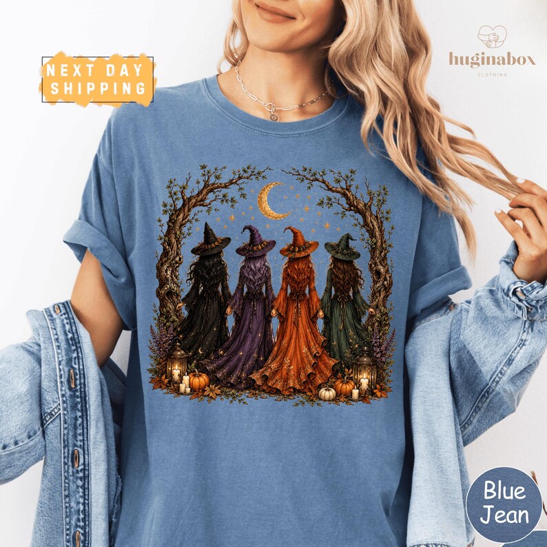 A woman wearing a blue t-shirt with a graphic design depicting four women dressed as witches standing in a circle under a crescent moon.